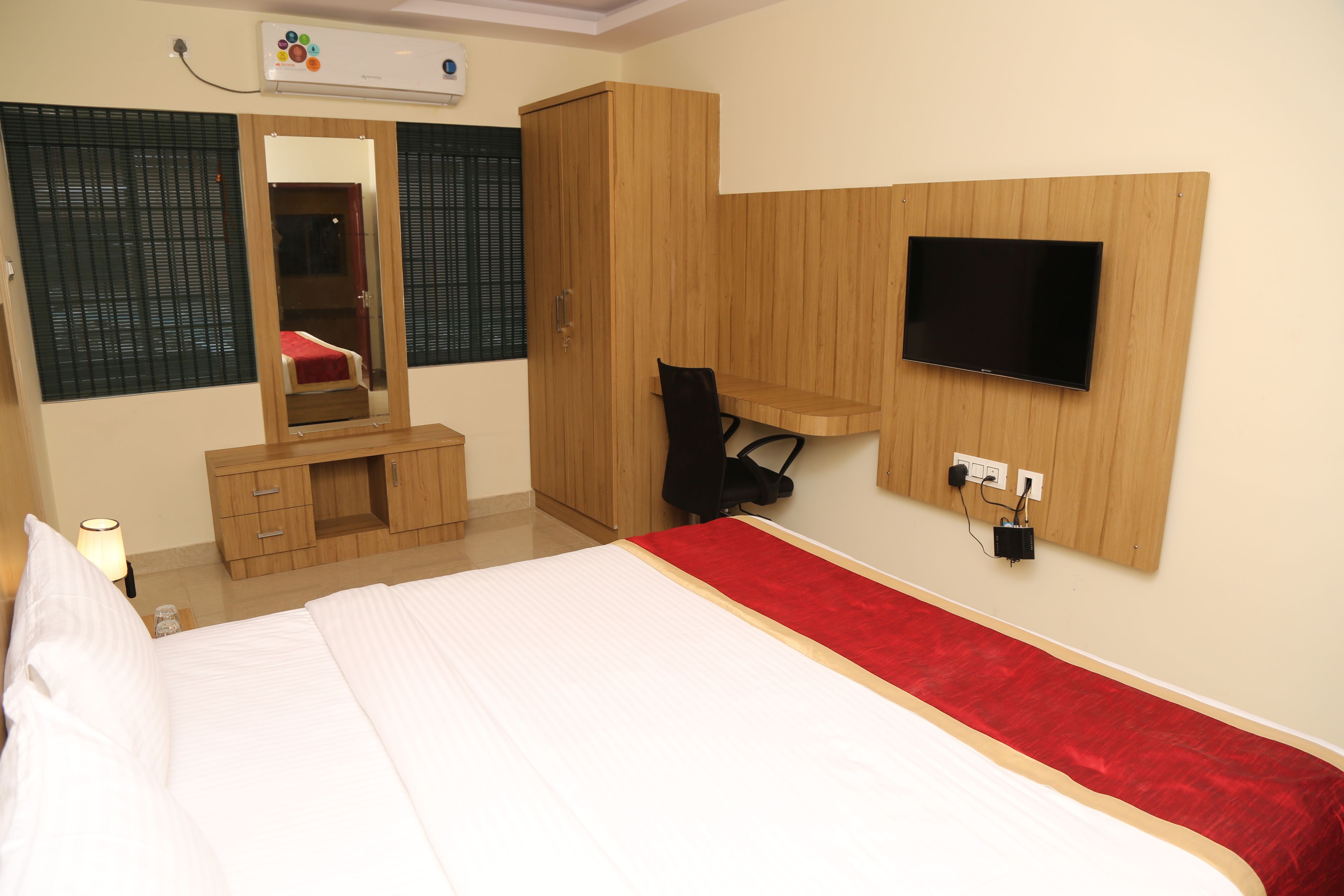 A bedroom with a bed, a wooden wardrobe, a desk with a chair, a TV mounted on the wall, and a mirror.