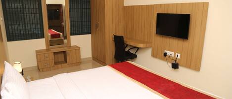 A bedroom with a bed, a wooden wardrobe, a desk with a chair, a TV mounted on the wall, and a mirror.