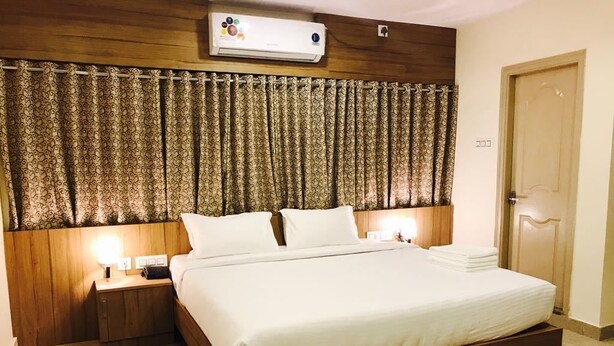 Una habitación de hotel con cabecera de madera, una cama grande con sábanas blancas, una mesita de noche con lámpara y una puerta con picaporte.