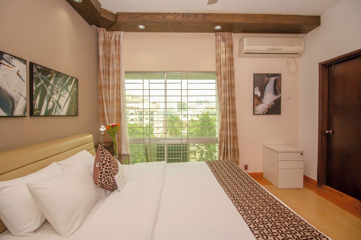 Habitación de hotel con cama, mesita de noche, unidad de aire acondicionado, ventana con cortinas y puerta.
