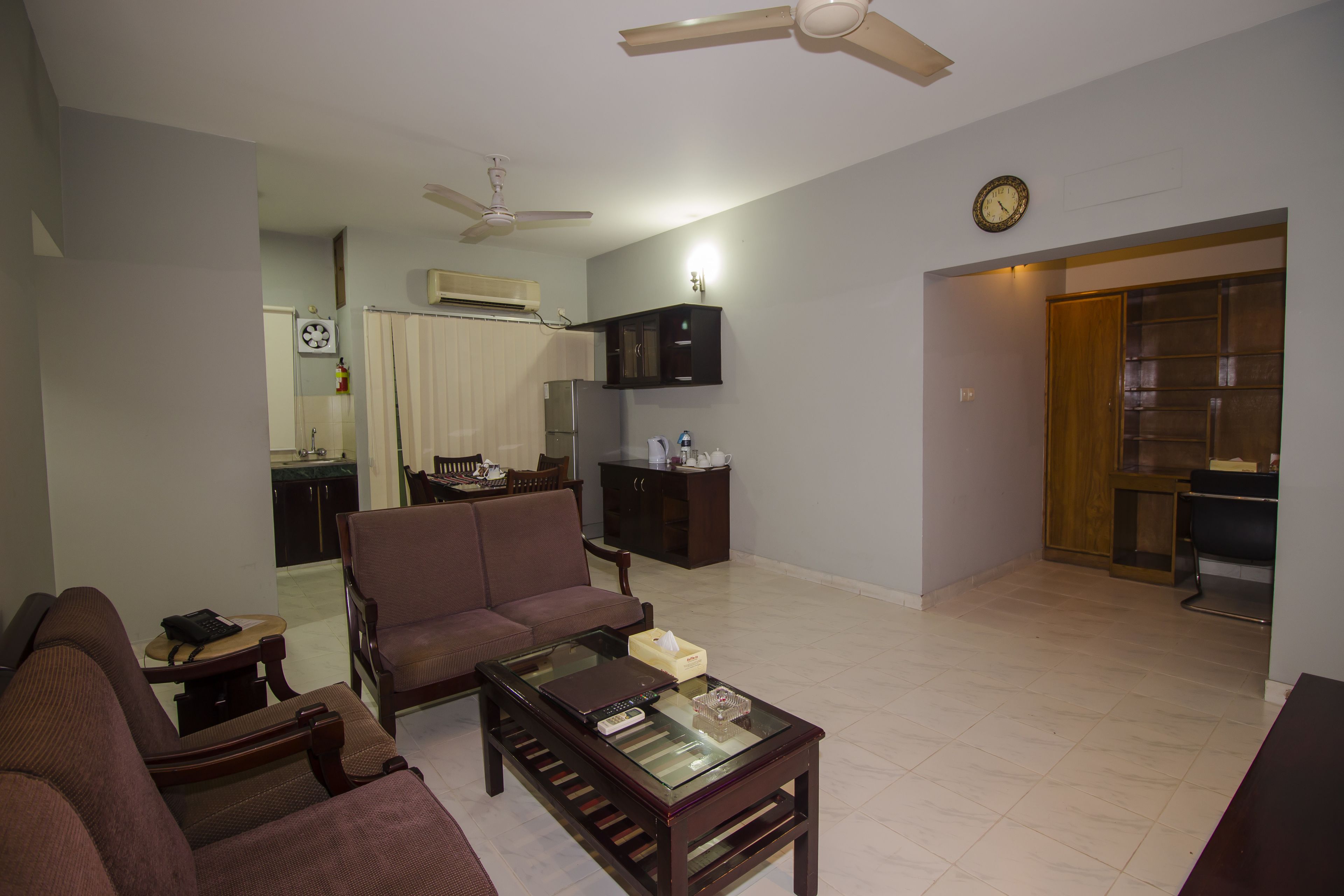 A living room with a sofa, coffee table, and a ceiling fan. There is a kitchen area in the background with cabinets and a refrigerator.