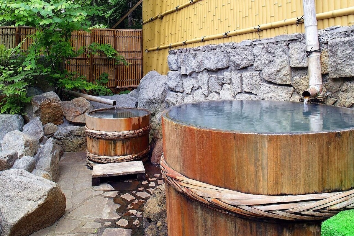 Un onsen japonais traditionnel, avec des baignoires en pierre et en bois, une clôture en bambou et un mur de pierre.