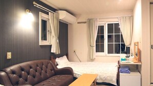 Habitación de hotel con un sofá de cuero marrón, dos camas individuales, una mesa de madera, un escritorio con una computadora y una ventana con cortinas.