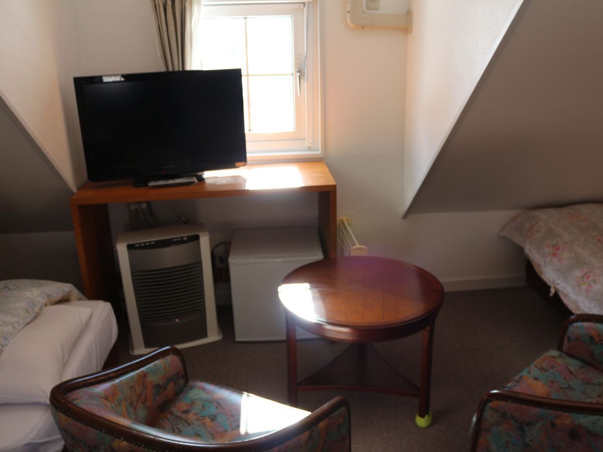 A small, compact room with a television on a wooden stand, a small refrigerator, a desk, a chair, and a bed with floral bedding.