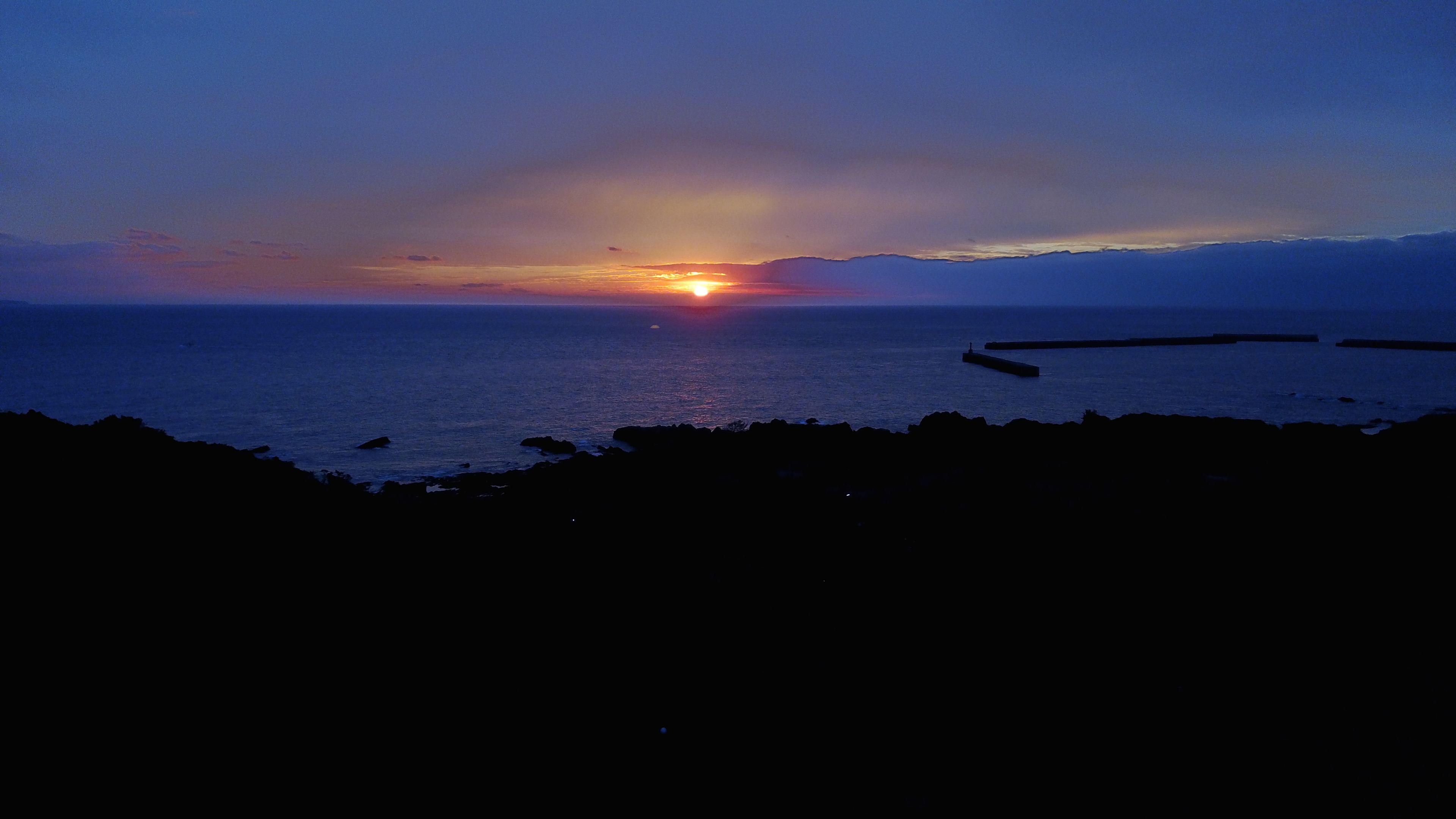 A coastal sunset with a rocky shoreline and a pier extending into the water.