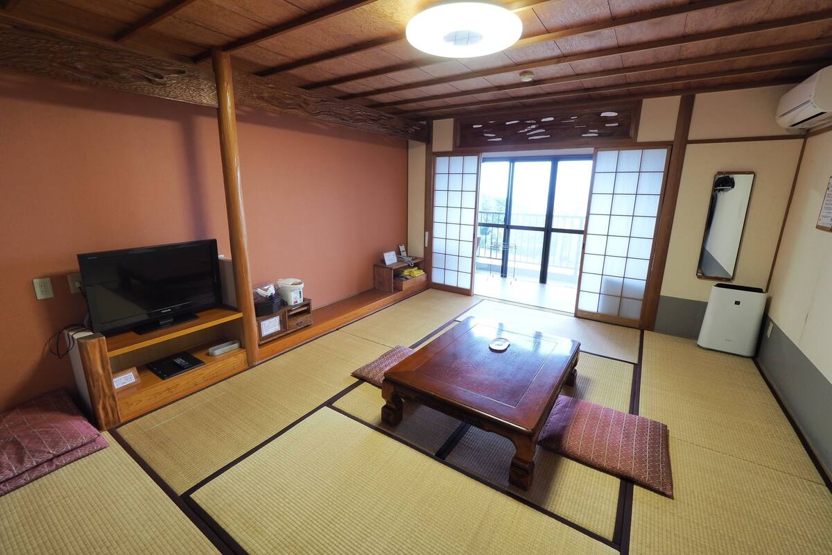 Une pièce de style japonais traditionnel, avec un sol recouvert de tatamis, un téléviseur posé sur un meuble en bois, une table avec un coussin et des portes coulissantes donnant sur un balcon.