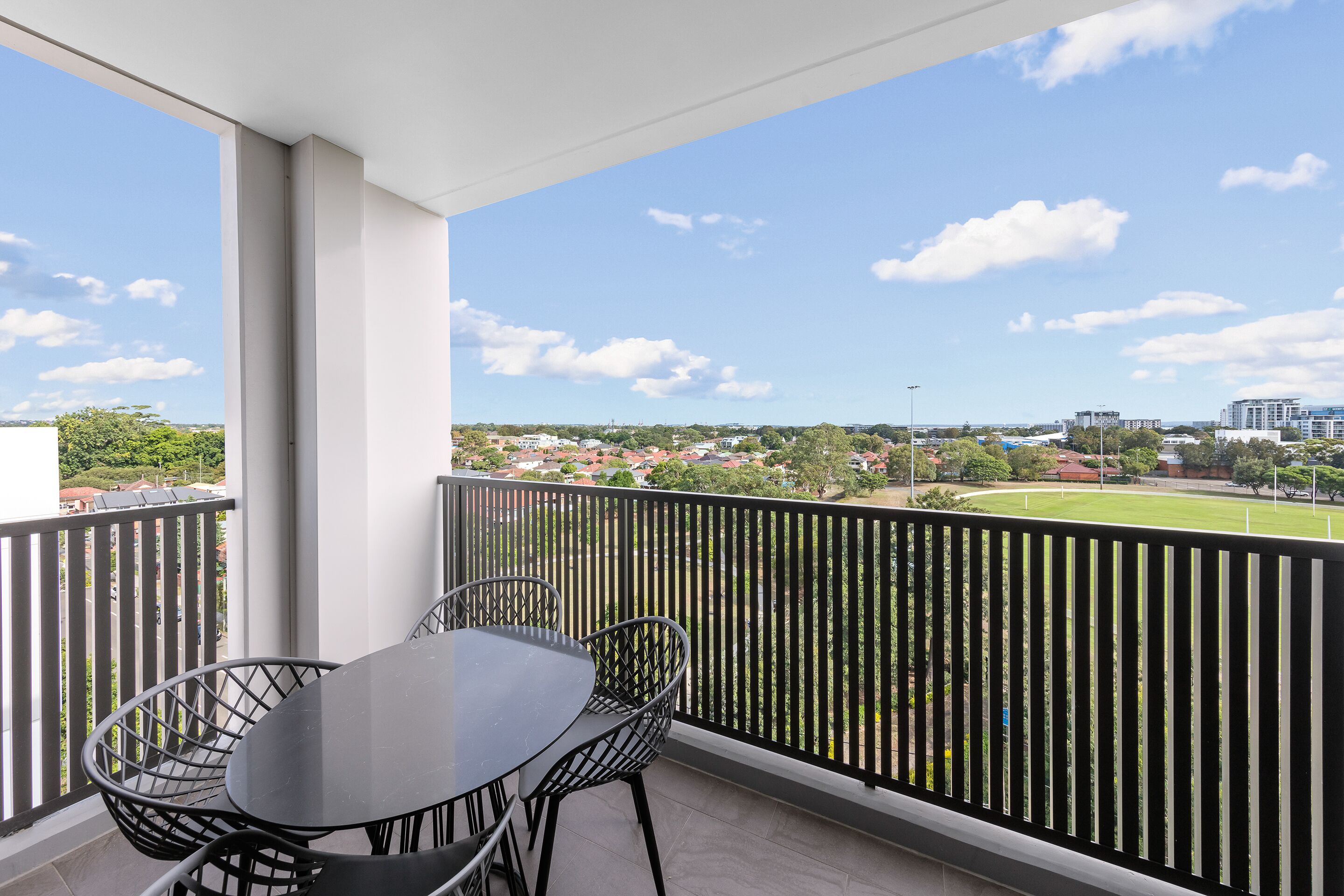 Uma varanda com mesa e cadeiras, com vista para um campo verde e edifícios ao longe.