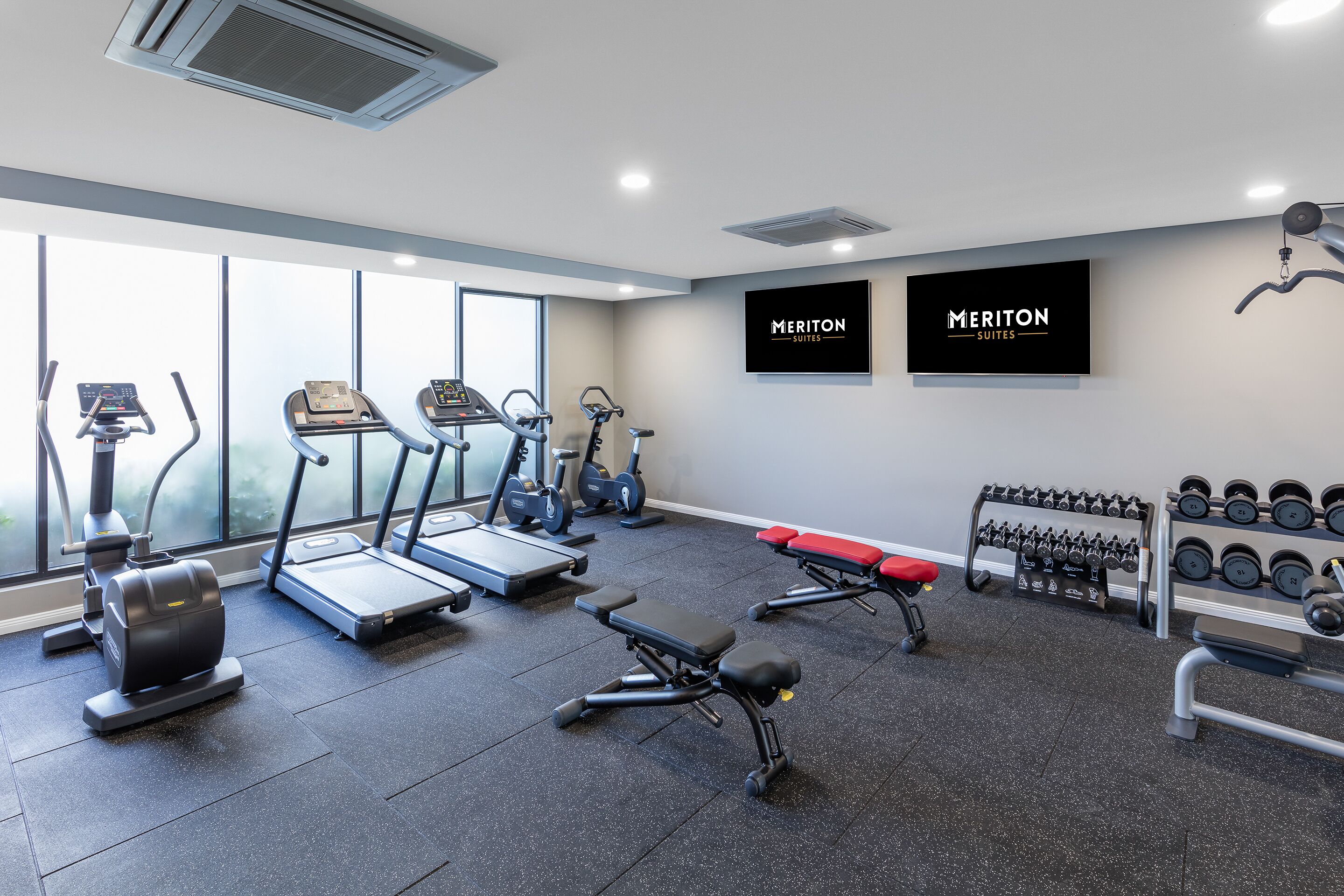 A modern gym with treadmills, elliptical machines, a bench, and a rack of dumbbells.