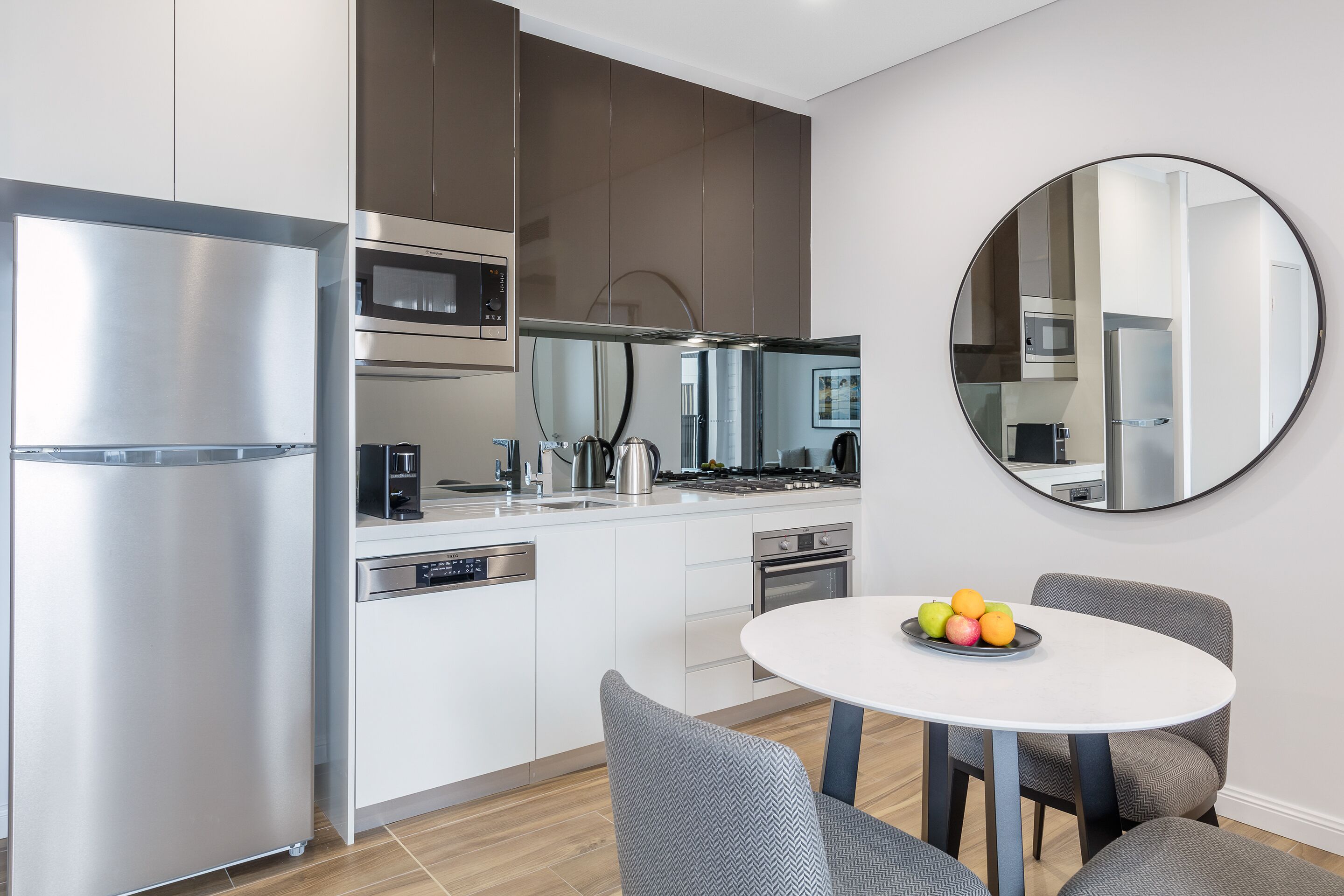 A modern kitchen with stainless steel appliances, a white dining table with a fruit bowl, and grey upholstered chairs.