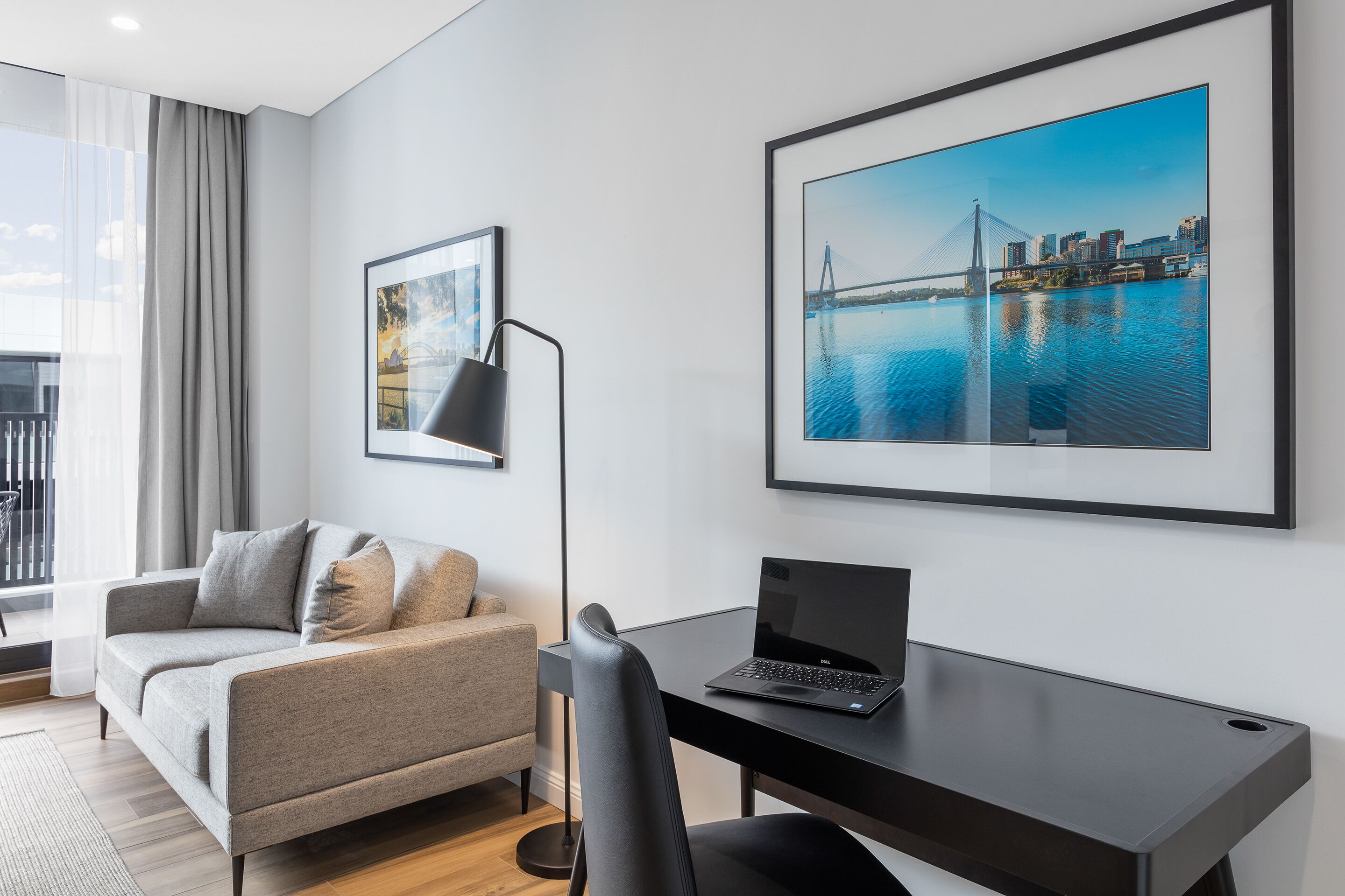 A modern hotel room with a grey sofa, a black desk with a laptop, and two framed pictures on the wall.