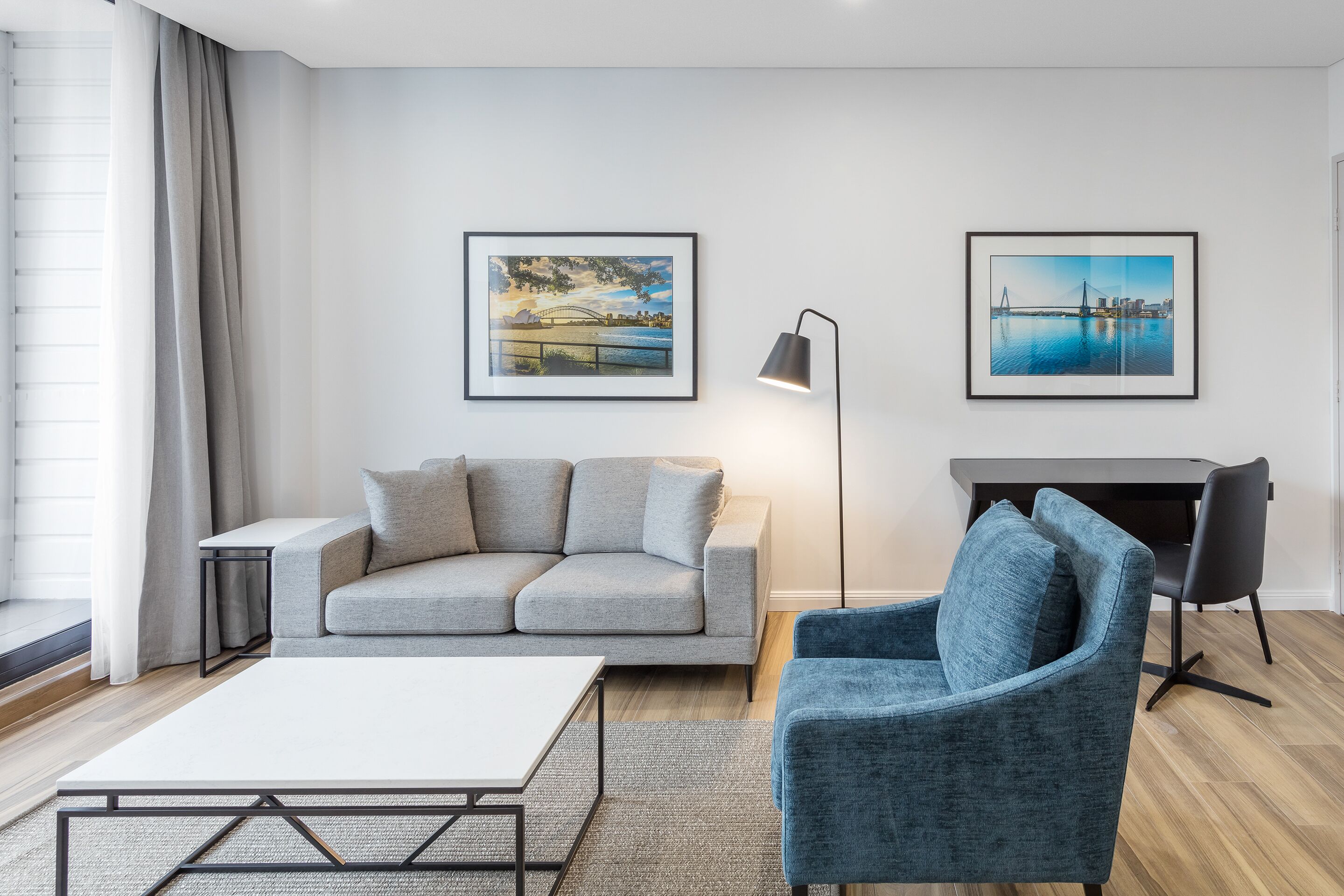 Ein modernes Wohnzimmer mit einem grauen Sofa, einem blauen Sessel, einem weißen Couchtisch, einer Stehlampe und zwei gerahmten Bildern an der Wand.