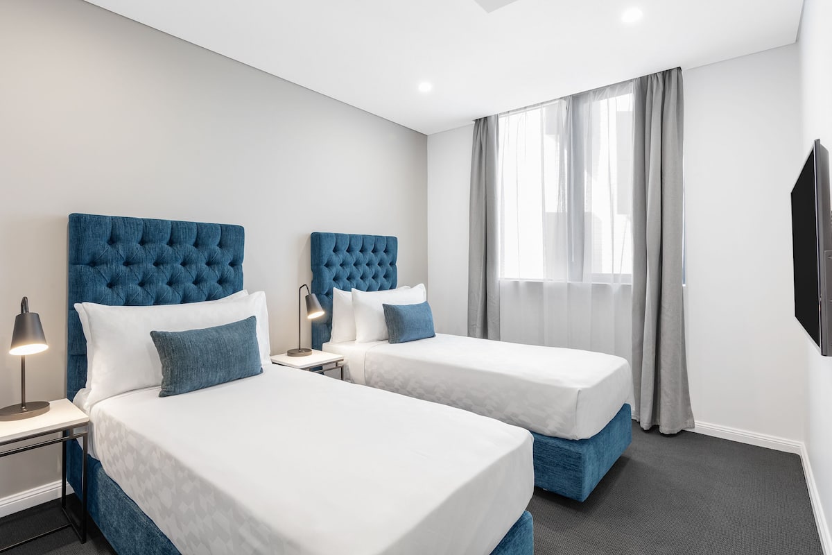 A hotel room with two beds, each with a blue headboard, white bedding, and a bedside lamp.