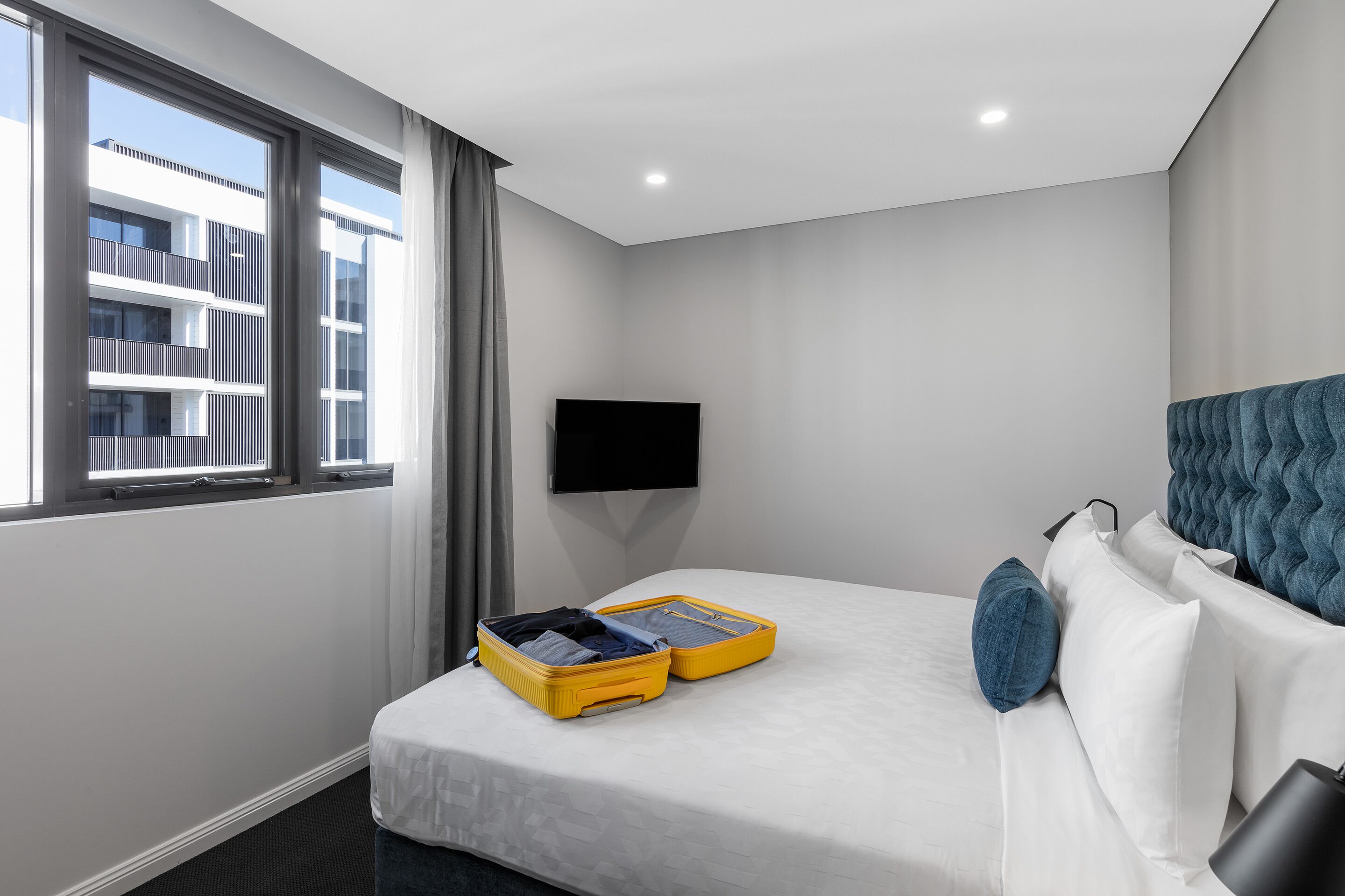 Habitación de hotel con una cama, un televisor de pantalla plana montado en la pared, una ventana con cortinas y una maleta sobre la cama.