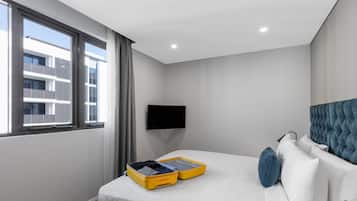 Habitación de hotel con una cama, un televisor de pantalla plana montado en la pared, una ventana con cortinas y una maleta sobre la cama.