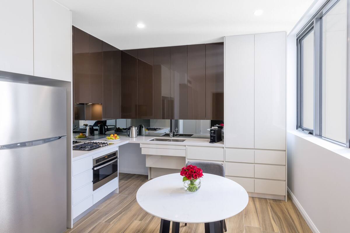 A modern kitchen with white cabinetry, a stainless steel refrigerator, and a dining area with a white table and vase of flowers.