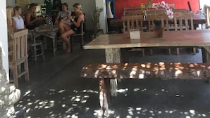 An outdoor seating area with wooden tables and chairs, a red wall with decorative patterns, and hanging woven light fixtures. - Padang Padang Breeze (Pecatu)