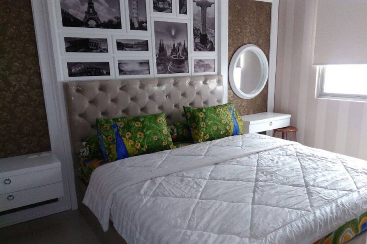 A bedroom with a tufted headboard, a bed with a white quilt, two green pillows with a peacock pattern, a round mirror on a bedside table, and a window with blinds.