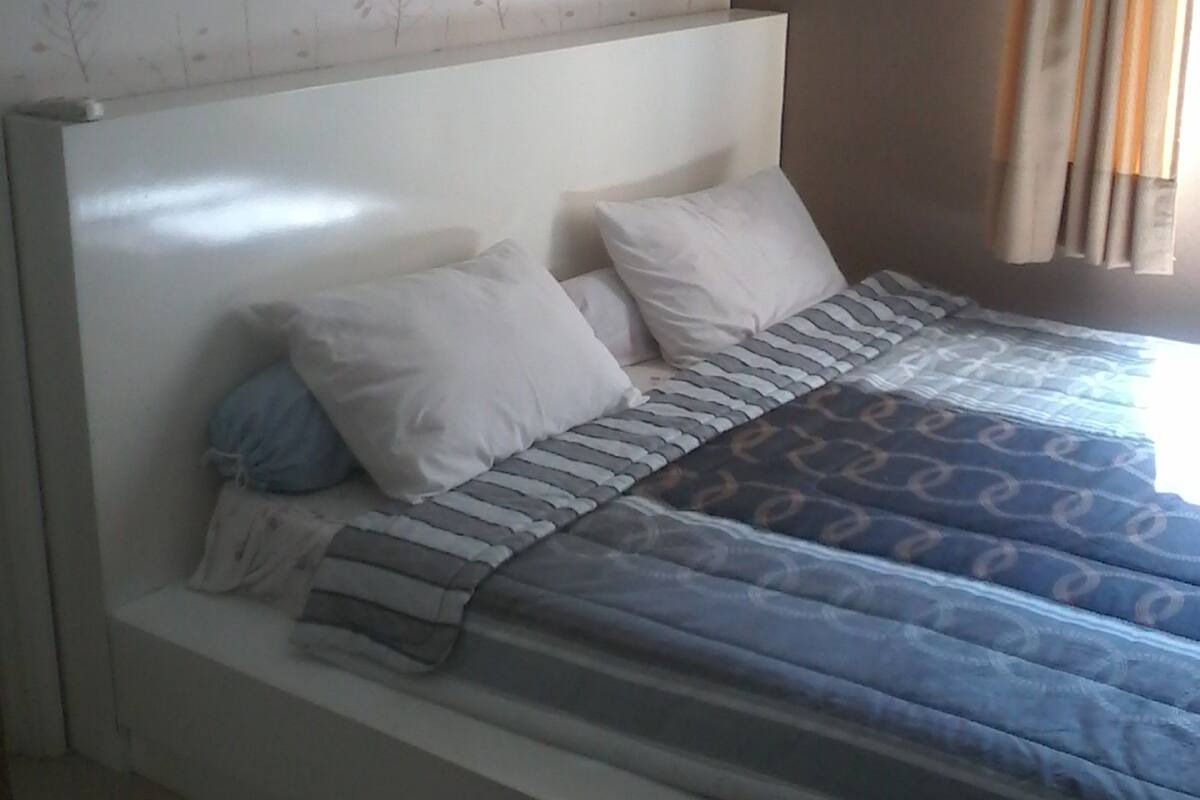 A bedroom with a bed, white headboard, patterned wallpaper, and a window with curtains.