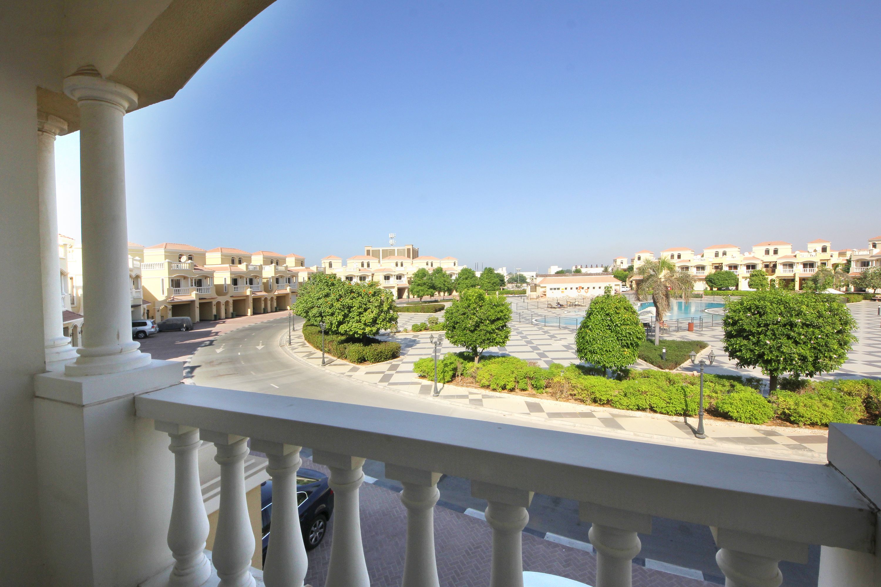 A balcony with white balustrades overlooking a courtyard with neatly arranged trees and a pool.