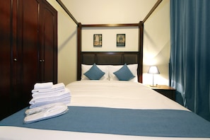 Ein Hotelzimmer mit einem ordentlich bezogenem Bett, einem Stapel Handtücher, einem Nachttisch mit Lampe und zwei gerahmten Bildern an der Wand.