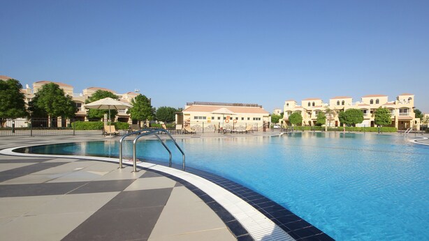 A swimming pool with a tiled deck, surrounded by buildings with balconies and trees. - Piks Key- Al Hamra Village (Ras Al Khaimah)