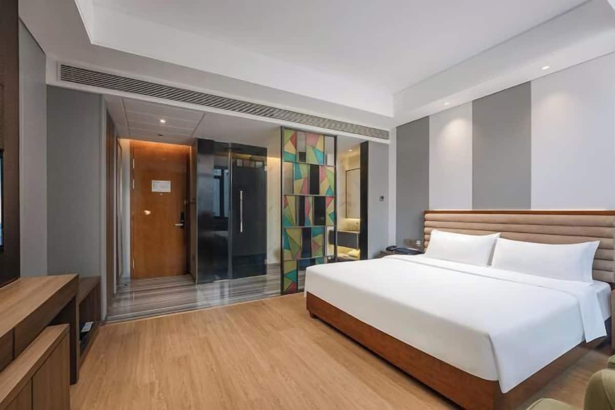 A modern hotel room with a large bed, a wooden bench, a desk, and a glass door leading to a bathroom.
