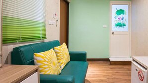 A compact room with a teal sofa, two yellow pillows, a wooden desk, and a small kitchenette. - Happy House (Liuqiu)