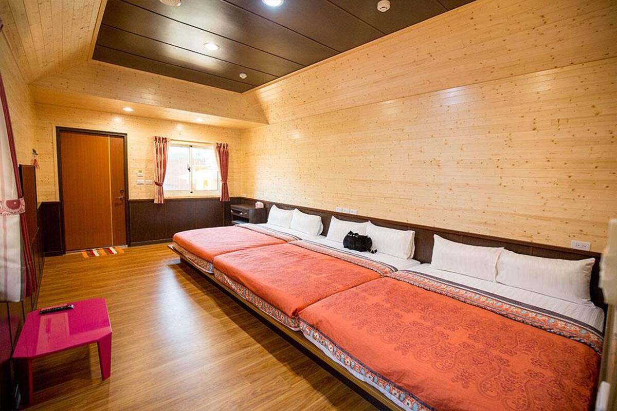 Una habitación con techo de madera, una cama con dos sábanas, una pequeña mesa rosa y una ventana con cortinas.