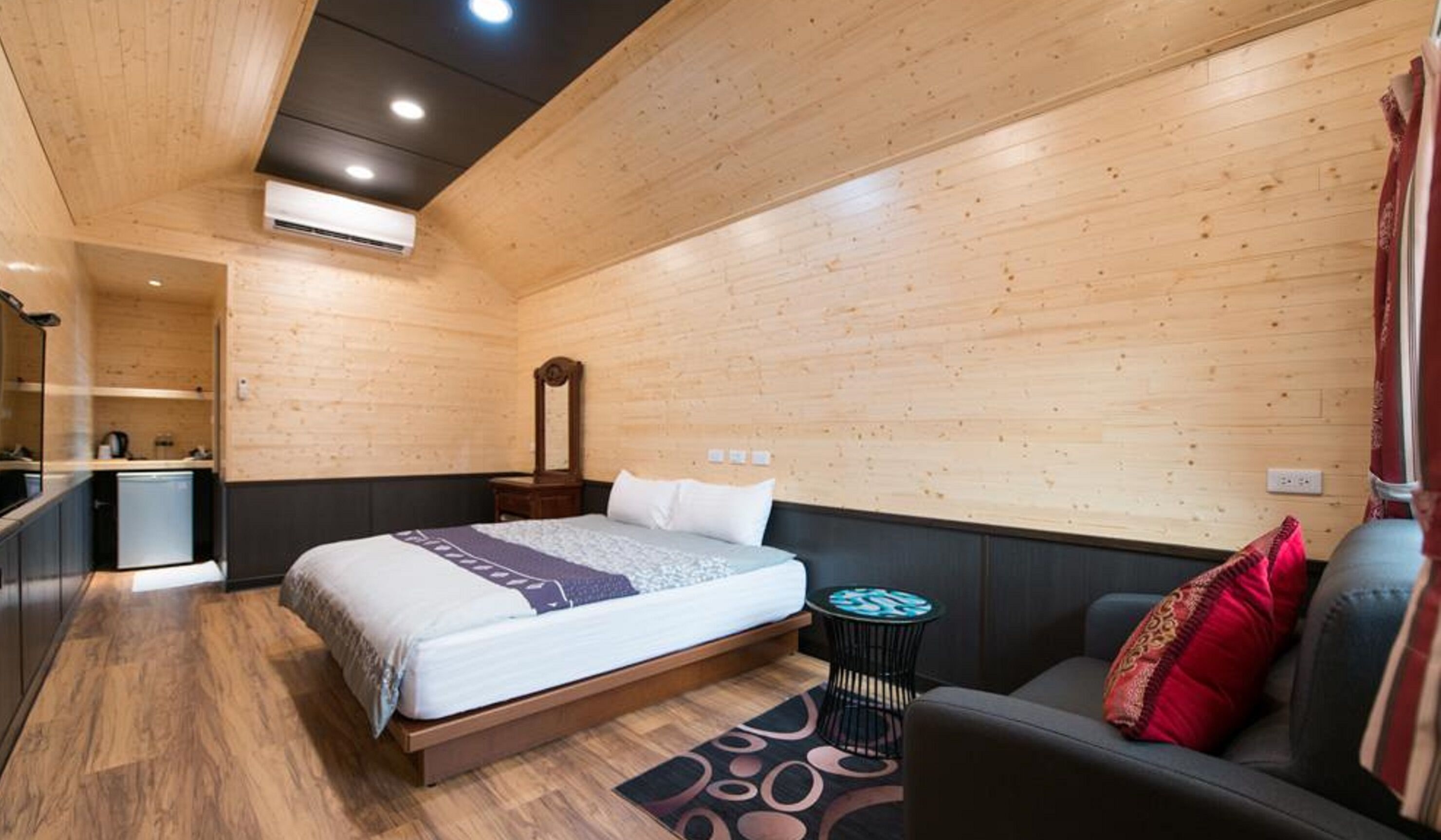 Un dormitorio moderno con techo de madera, una cama con colcha estampada, una mesita de noche, un espejo, una pequeña mesa redonda con una silla y un sofá con un cojín rojo.