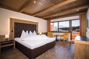 Habitación de hotel con una cama grande, mesitas de noche, un escritorio y una silla. Hay una ventana con vistas a las montañas.