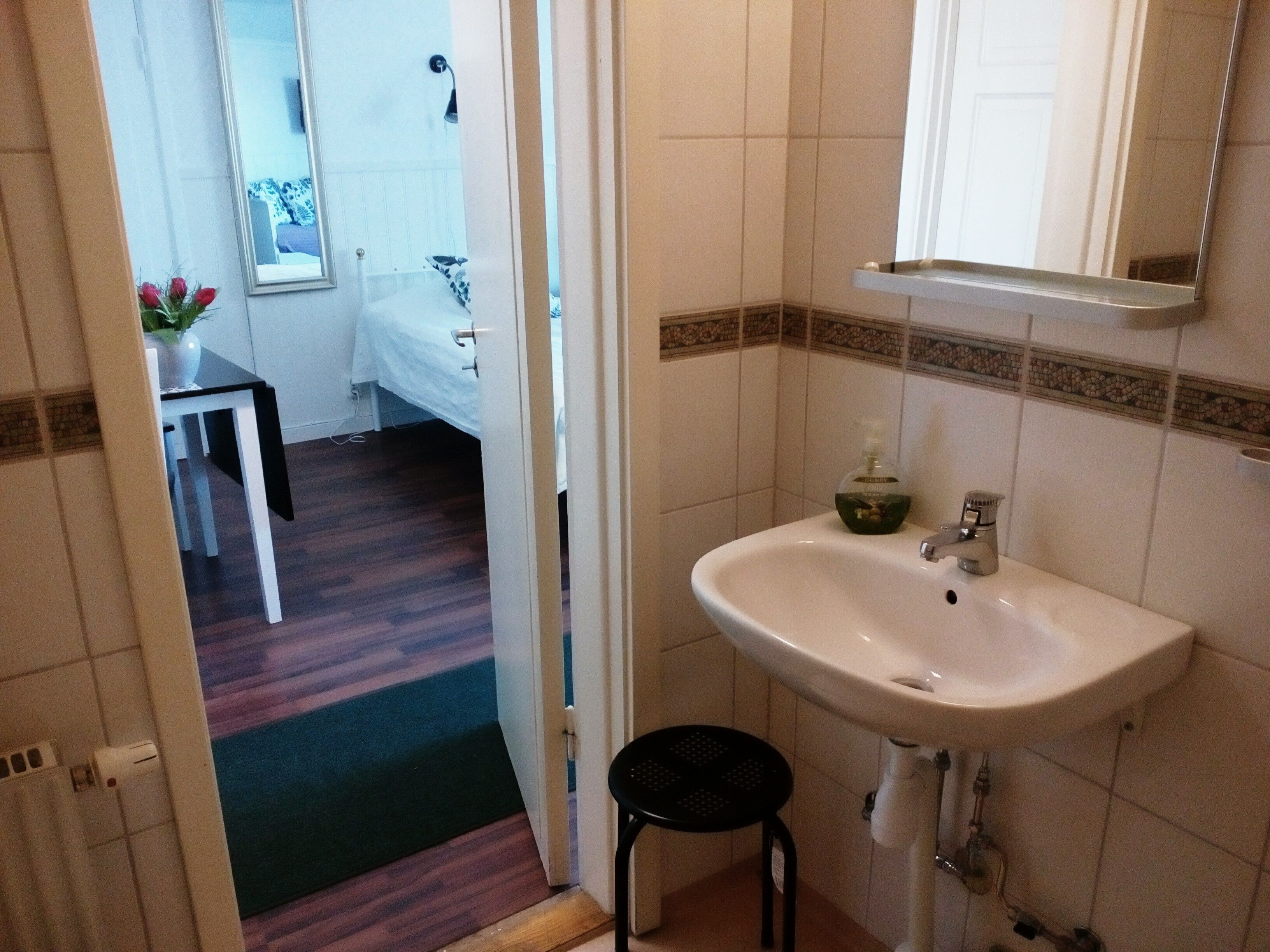 Un baño con un lavamanos blanco, un espejo encima y un taburete pequeño. Hay una puerta que da a un dormitorio con una cama y una mesita de noche.