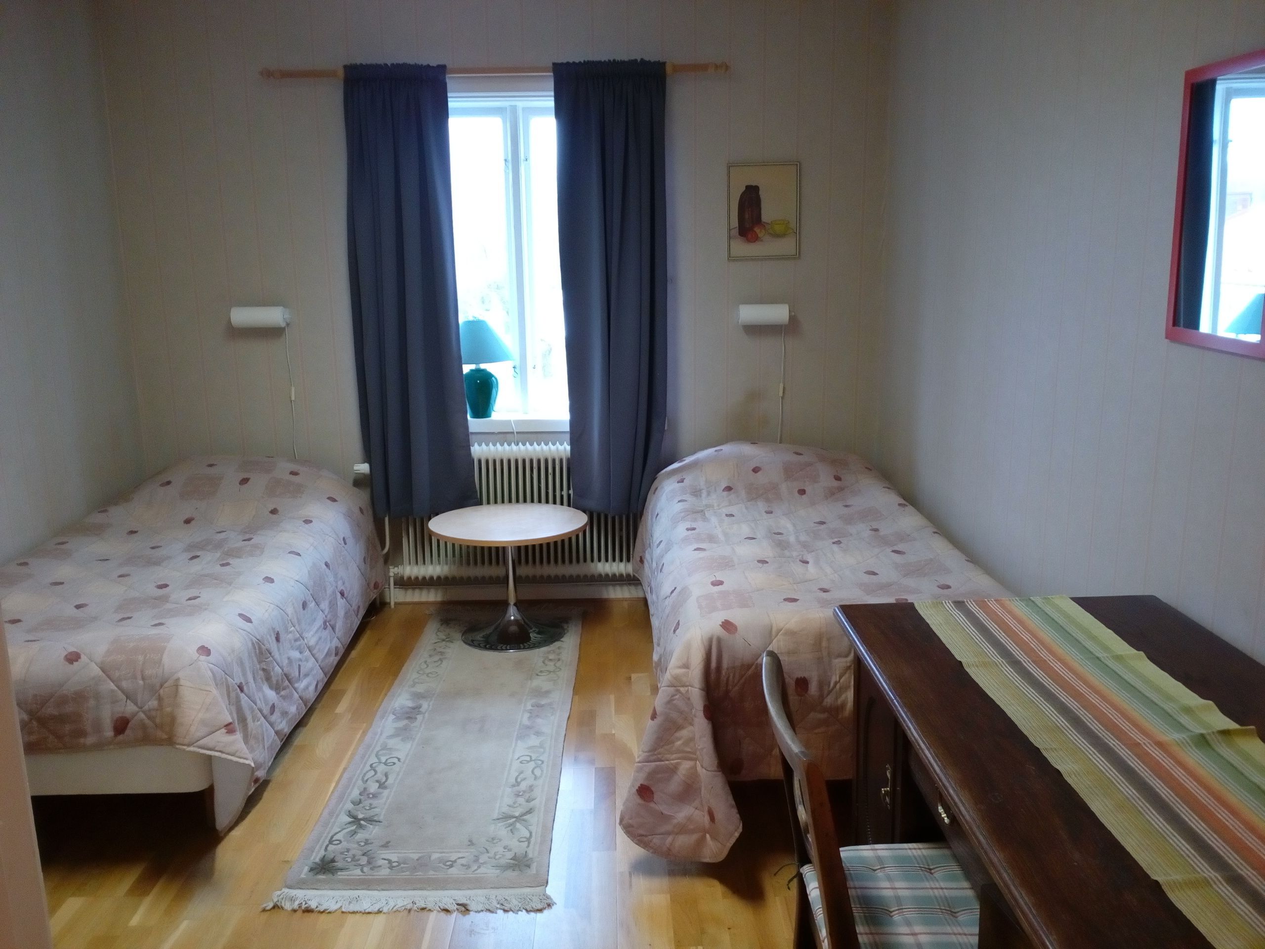 A small, well-lit room with two single beds, a small round table, and a window with curtains.