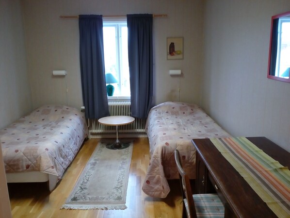Una habitación pequeña y bien iluminada con dos camas individuales, una mesita redonda y una ventana con cortinas.