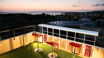 A rooftop terrace with two red bar stools, a glass railing, and a view of the cityscape and mountains in the distance.