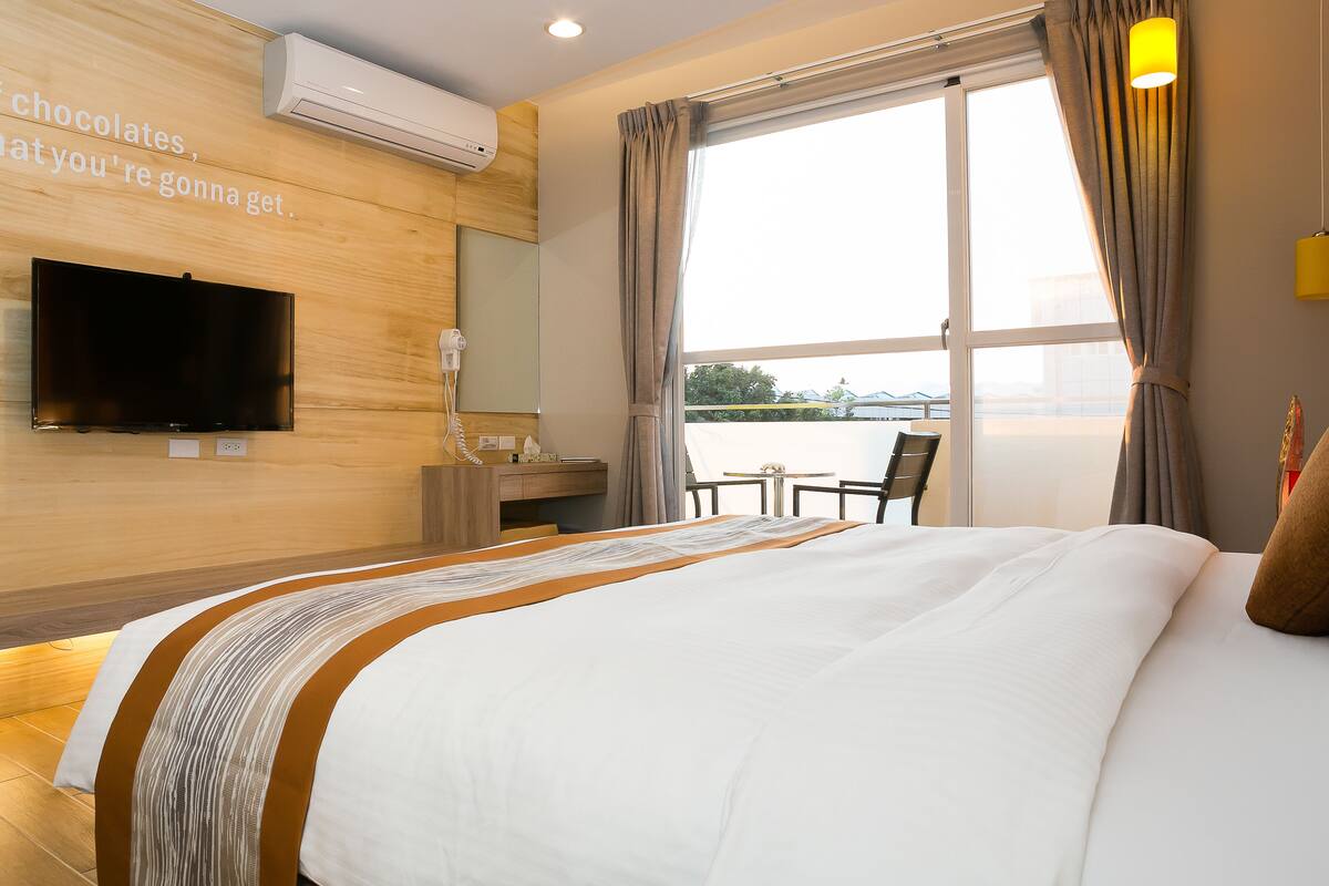 A hotel room with a large bed, a television mounted on the wall, a desk with a chair, and a window with curtains.