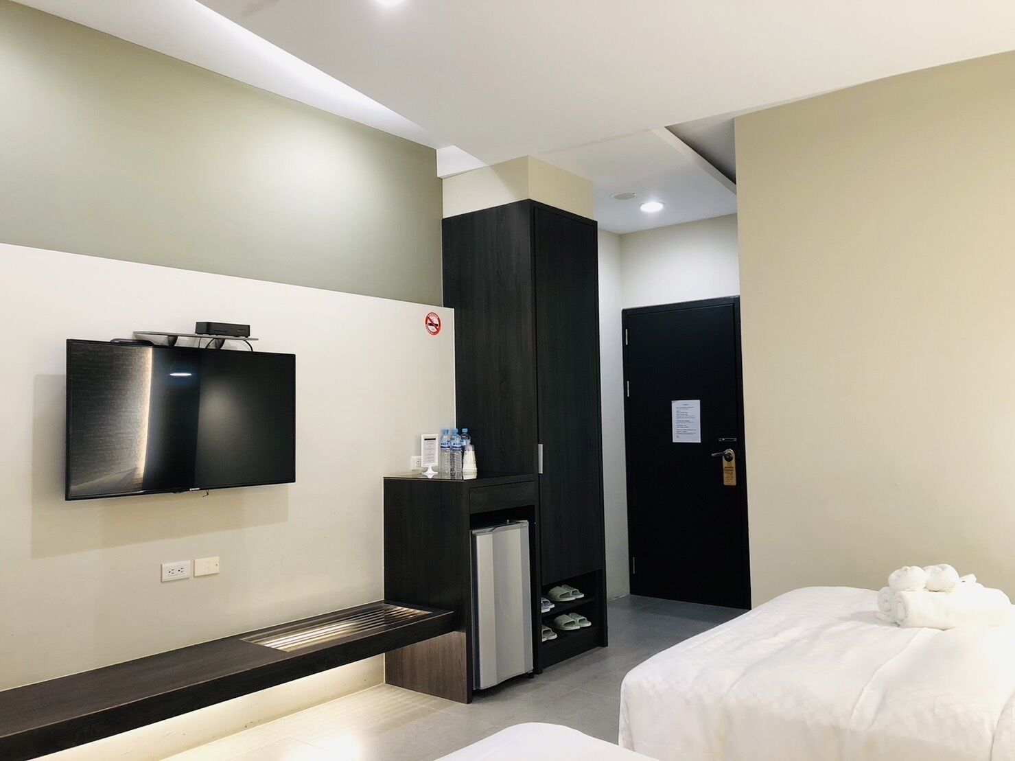 A hotel room with a flat-screen TV mounted on the wall, a small refrigerator, a shoe rack, a bed with white bedding, and a door with a sign.