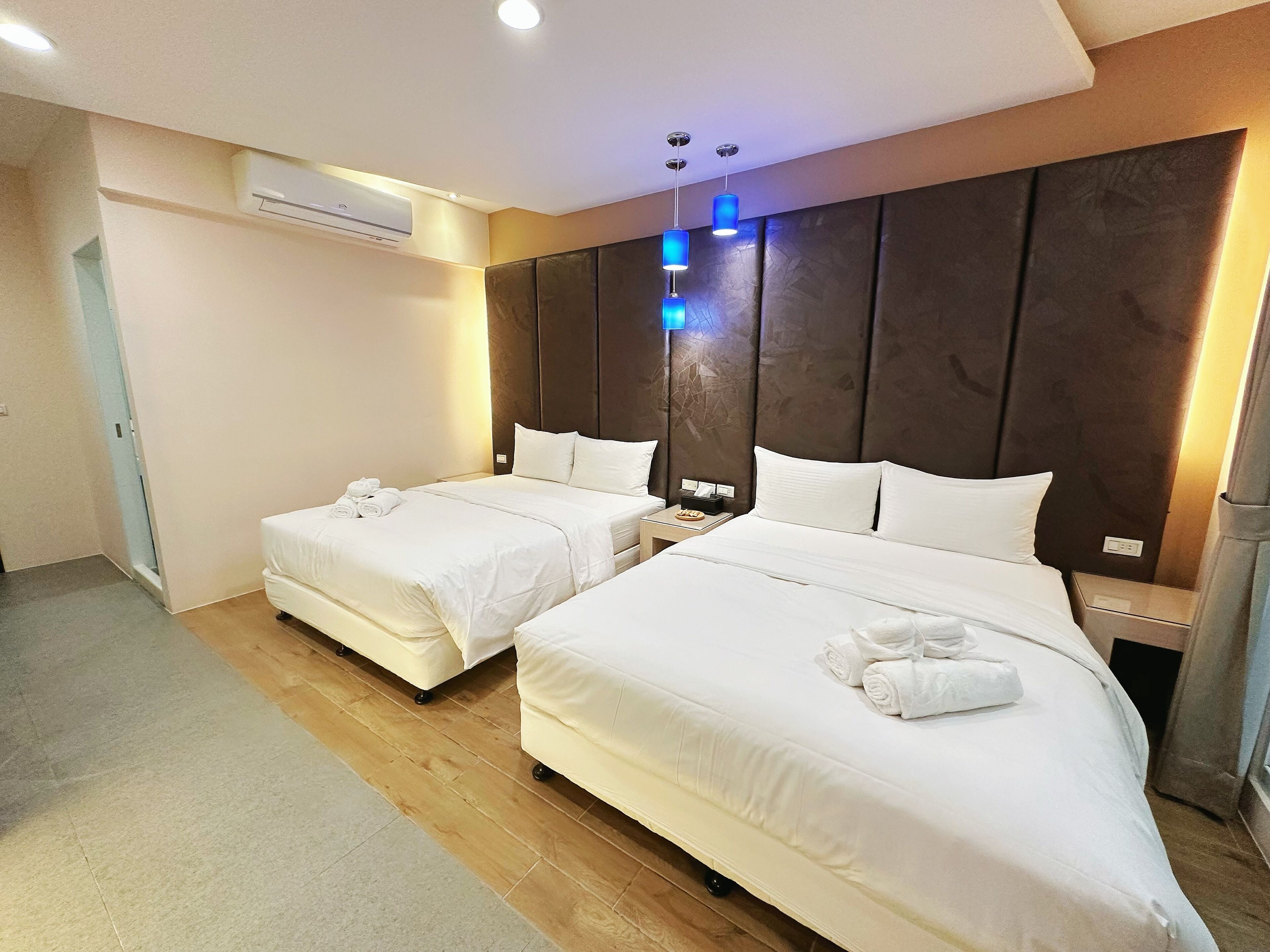 A hotel room with two beds, a wooden floor, an air conditioning unit, and a nightstand with a lamp.