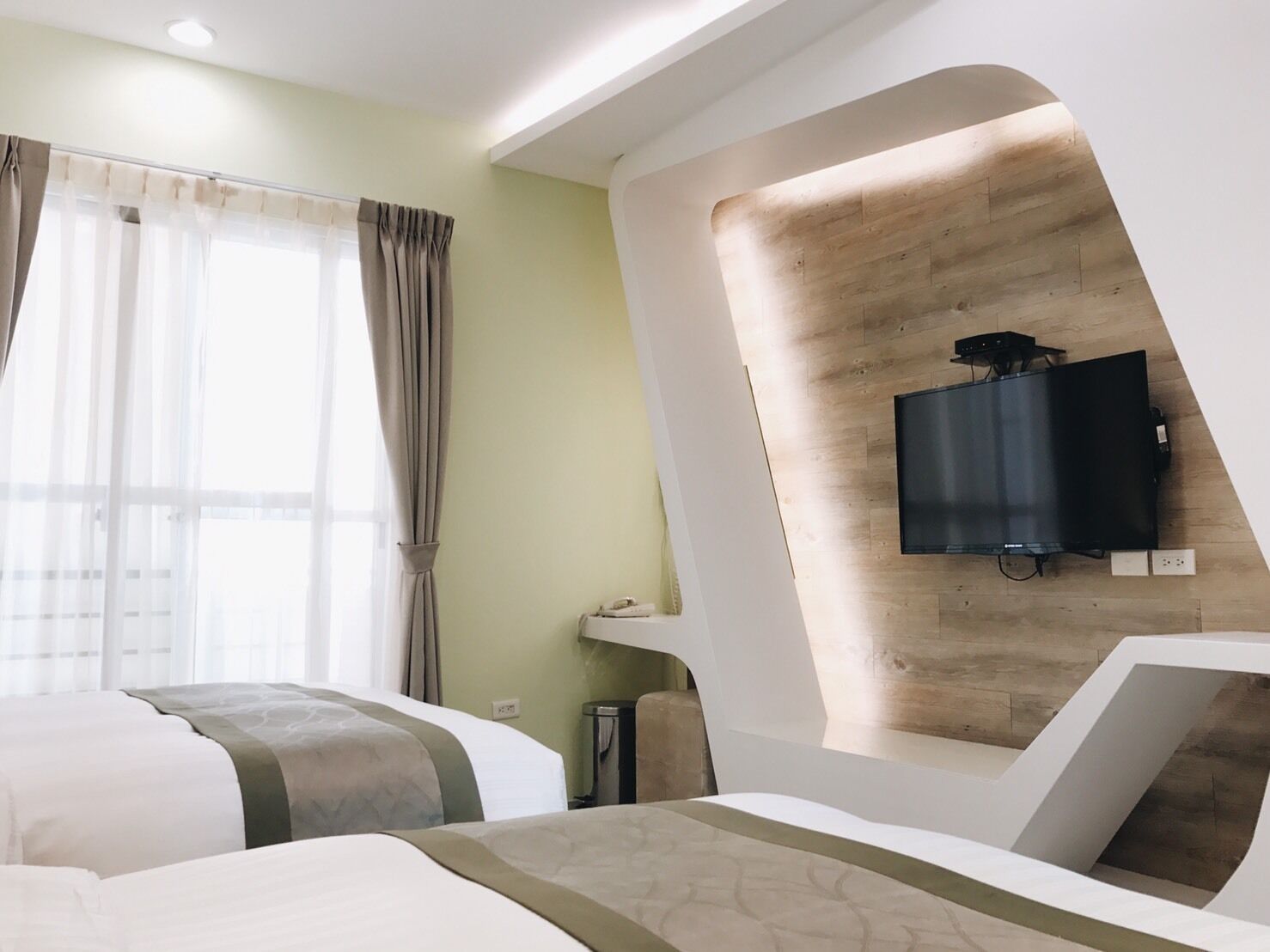 A modern hotel room with a large bed, a TV mounted on a wooden wall, and a window with curtains.