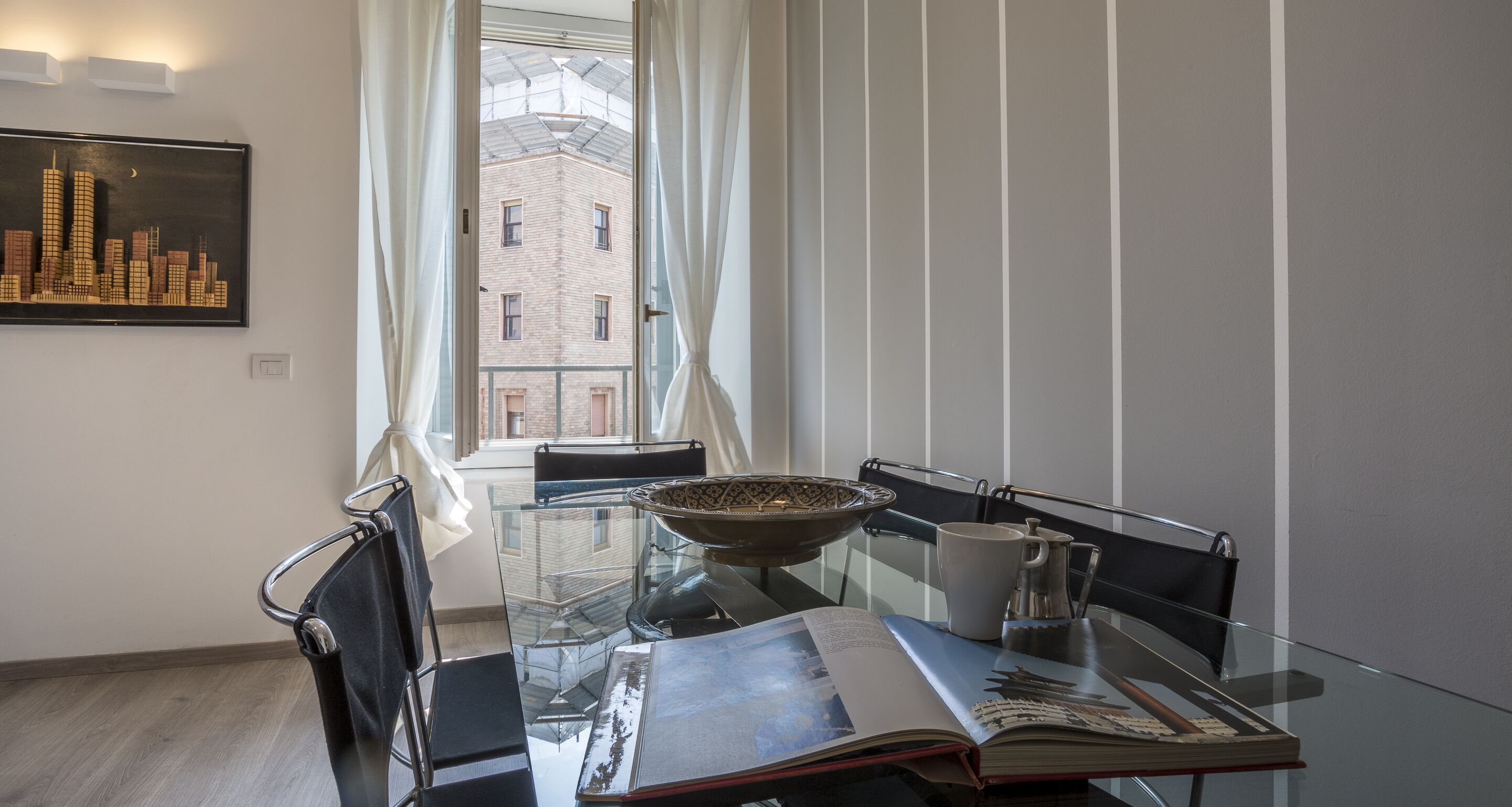 A modern dining area with a glass table, metal chairs, a bowl, a mug, and a book.