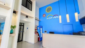 A modern hotel lobby with a blue wall featuring the 'HOP INN' logo, a reception desk, a vending machine, and a potted plant. - Hop Inn Nakhon Sawan (Nakhon Sawan)