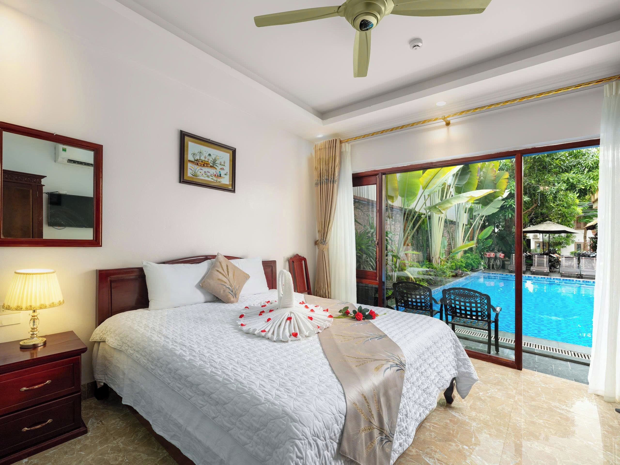 Habitación de hotel con cama, mesita de noche, lámpara, ventilador de techo y un ventanal con vistas a una piscina y vegetación.