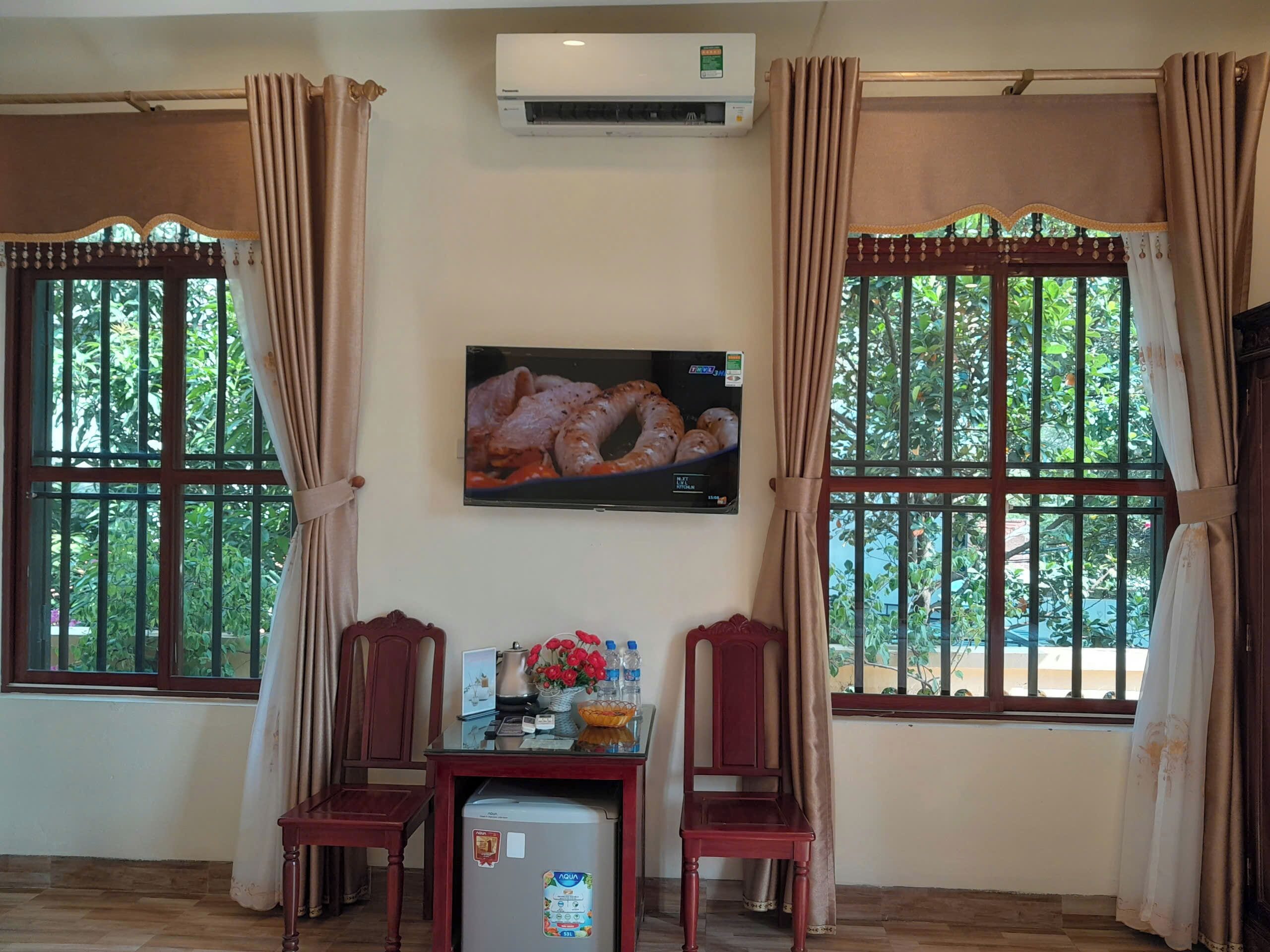 Quarto com duas cadeiras de madeira, uma pequena mesa com chaleira e flores, um refrigerador e uma televisão fixada na parede.
