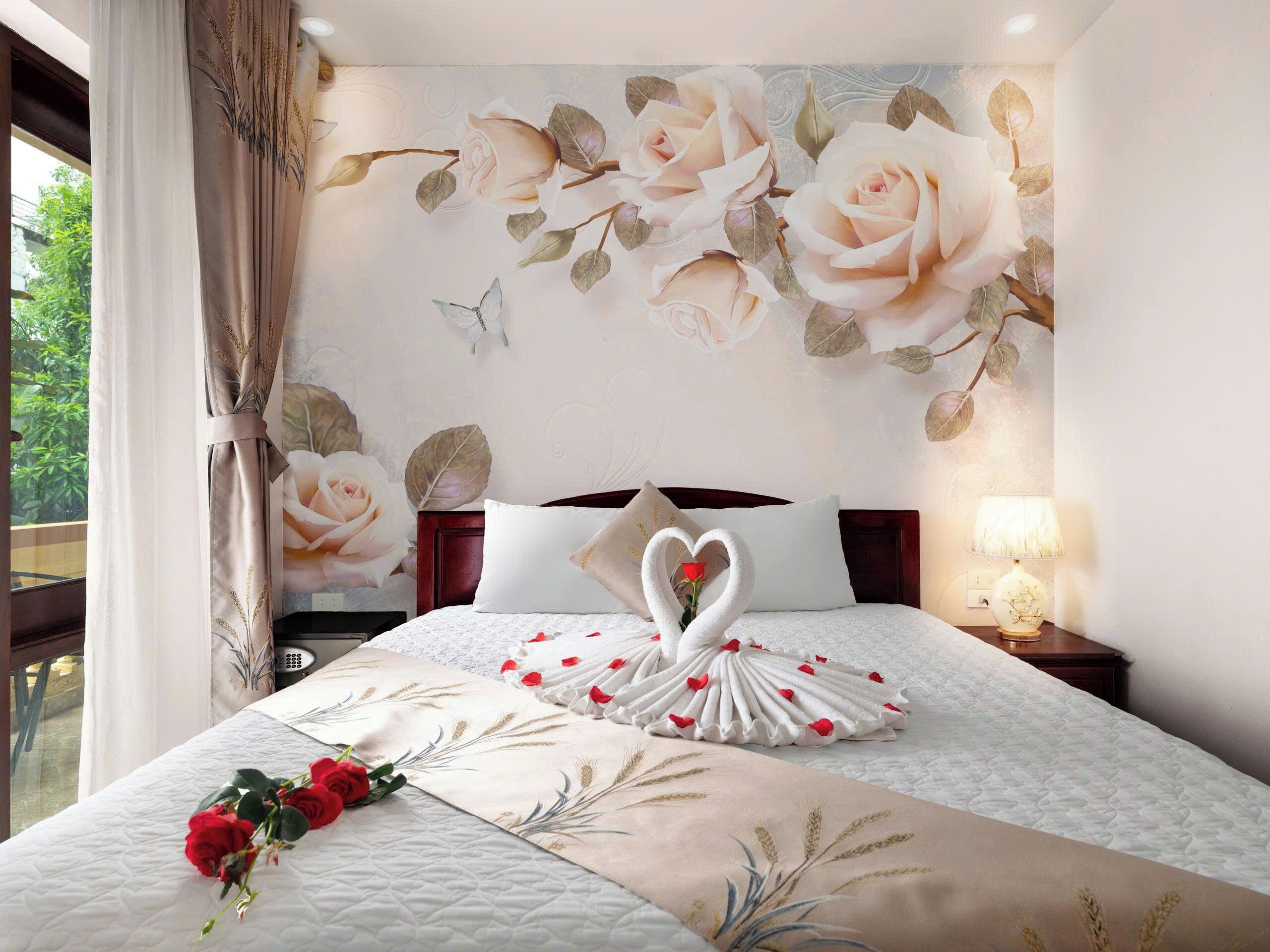 A neatly made bed with a floral bedspread, a heart-shaped towel arrangement, and a bouquet of roses.