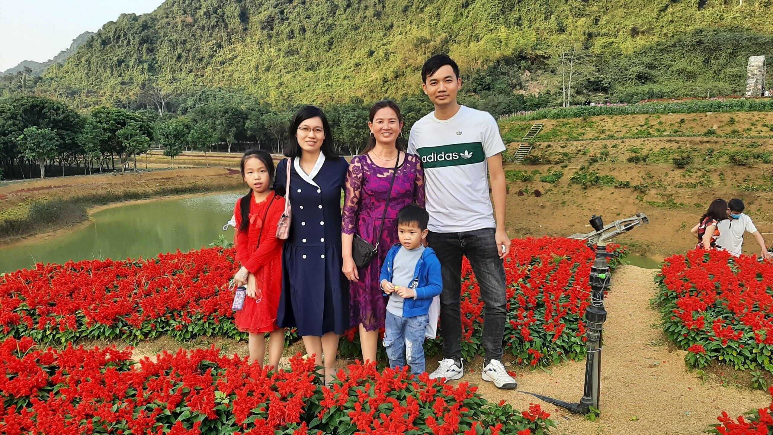 A group of people standing in a garden with red flowers and a pond in the background.