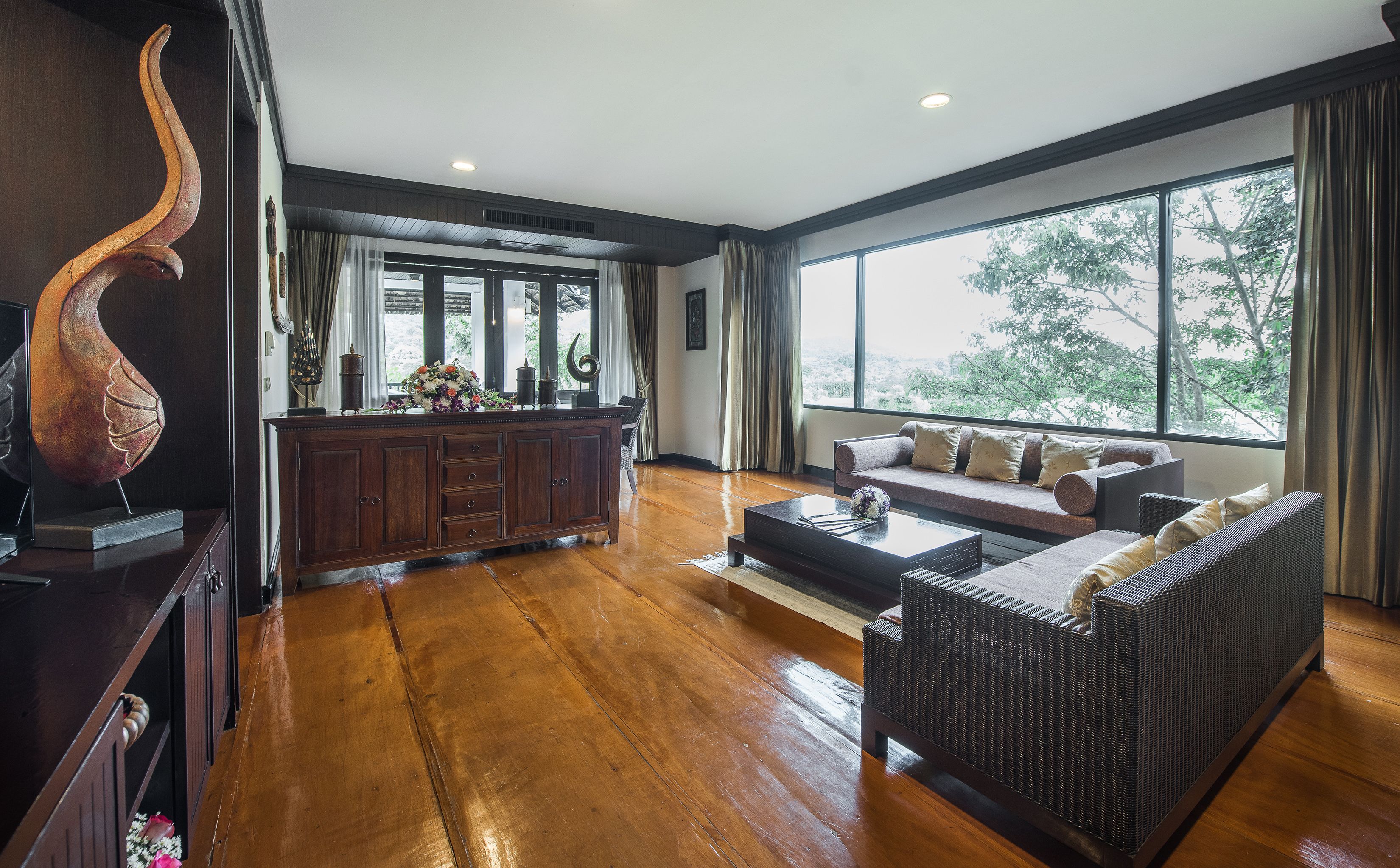 Amplia sala de estar con piso de madera, un conjunto de sofás, una mesa de comedor con un arreglo floral y un ventanal con vista a árboles.