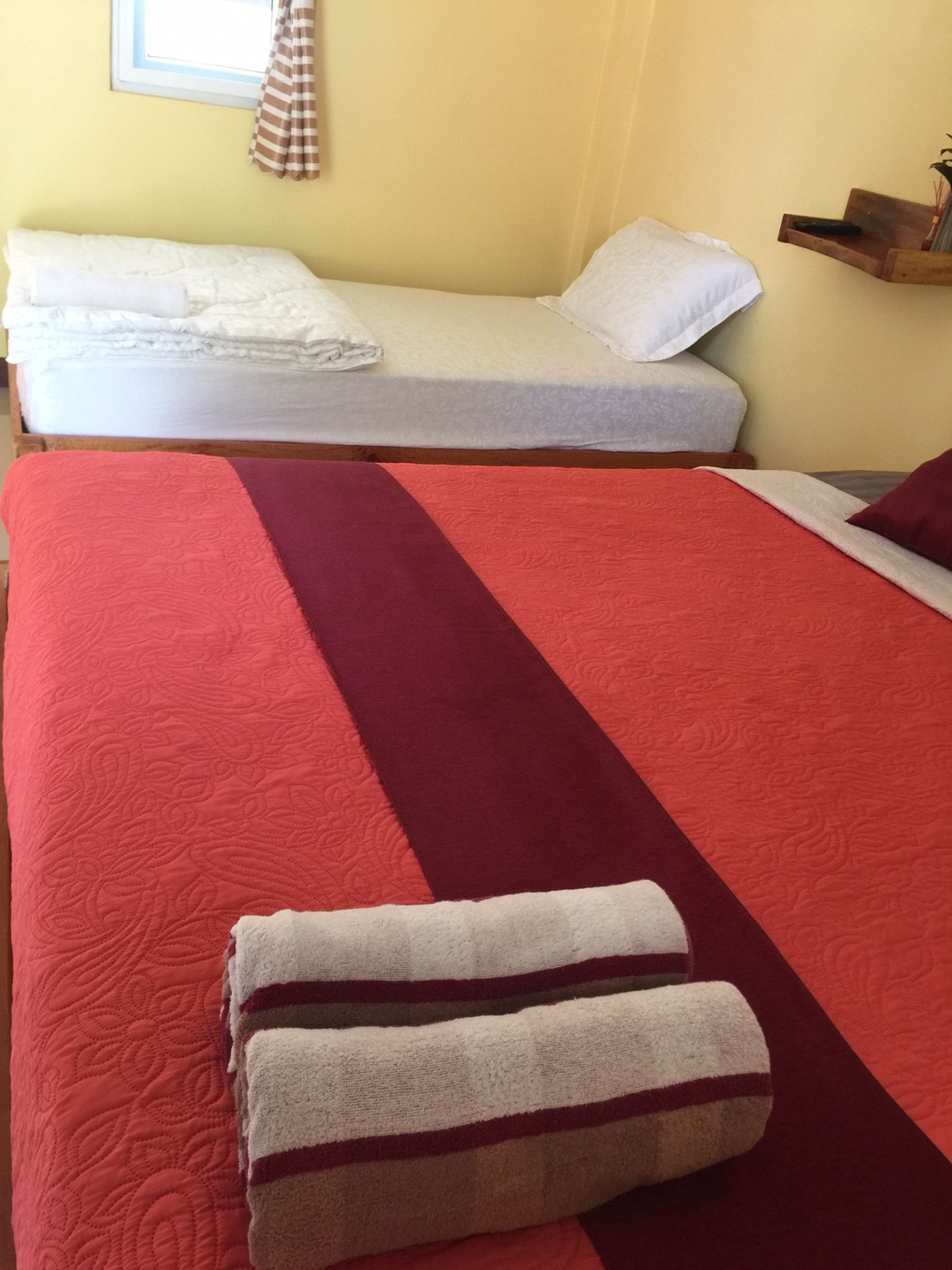 Una habitación con dos camas, una con colcha roja y blanca y la otra con colcha blanca. Hay una estantería de madera con una planta y un control remoto.