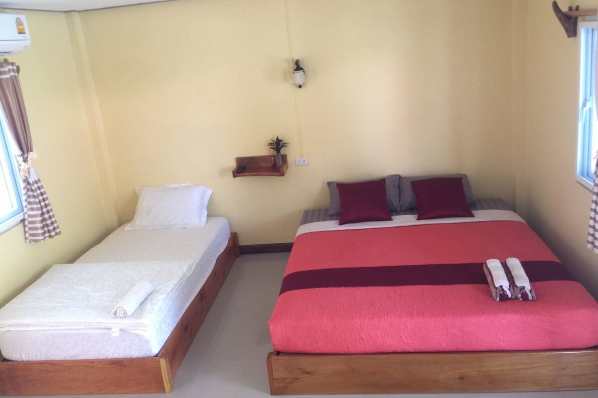 Ein Zimmer mit zwei Betten, eines mit einer weißen Matratze und das andere mit einer roten. An einem kleinen Regal stehen eine Pflanze und ein Telefon.