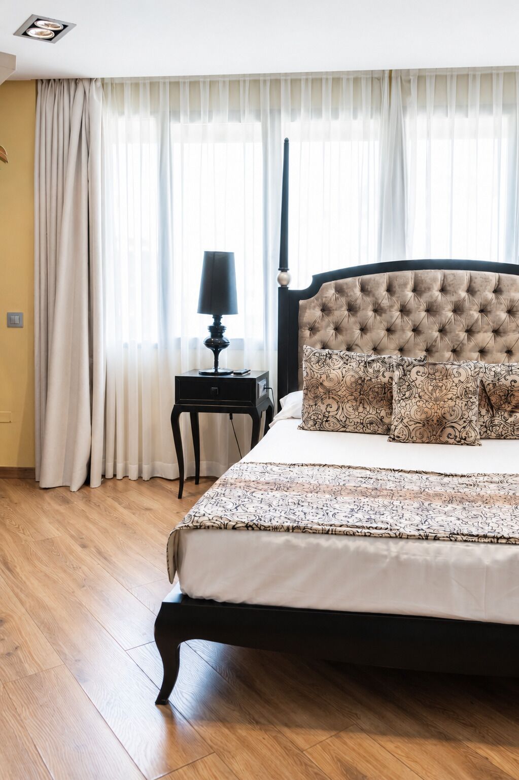 Ein Schlafzimmer mit einem großen Bett, einem Nachttisch mit Lampe und einem Fenster mit durchsichtigen Vorhängen.
