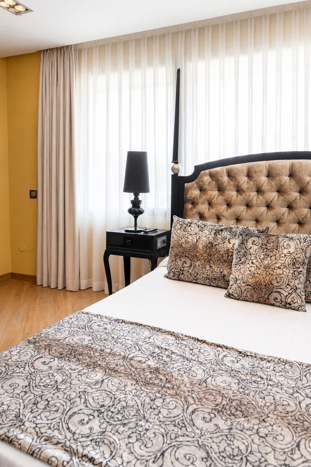 A hotel room with a large bed, a nightstand, a lamp, and sheer curtains.