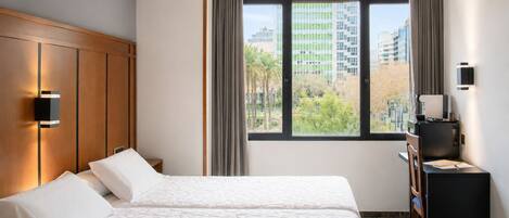 A hotel room with two beds, a desk with a chair, a window with curtains, and a view of buildings.